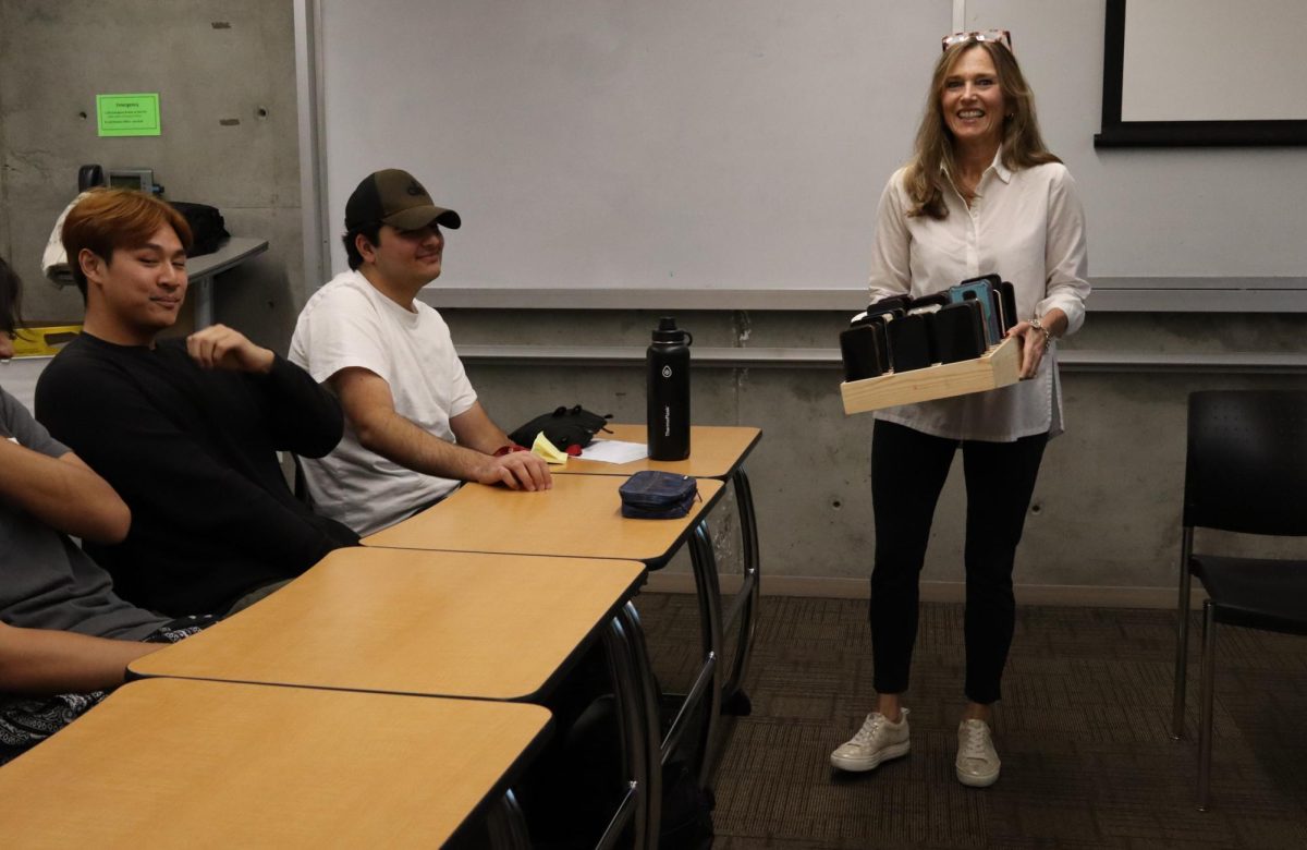 A woman smiling while holding a tray of phones engages with students in a classroom setting
